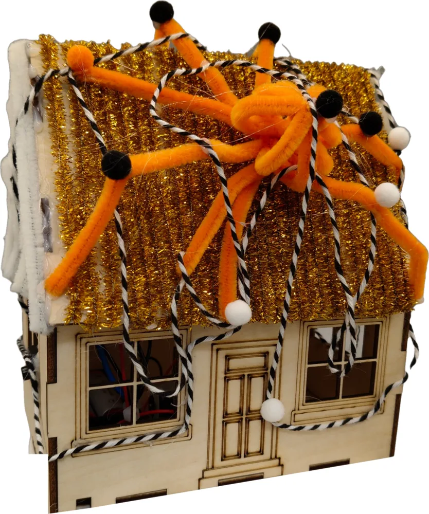 A laser-cut wooden haunted house with a tinsel roof and an orange pipe-cleaner spider